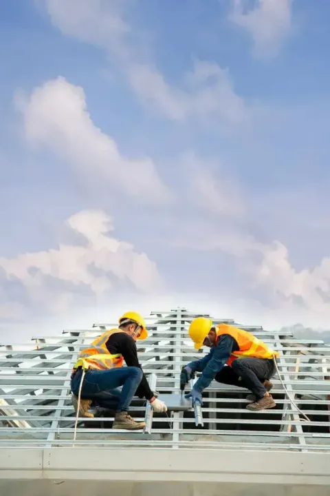 Two workers in yellow hard hats and safety vests from a local roofing company in Aurora, IL install a metal roof frame.