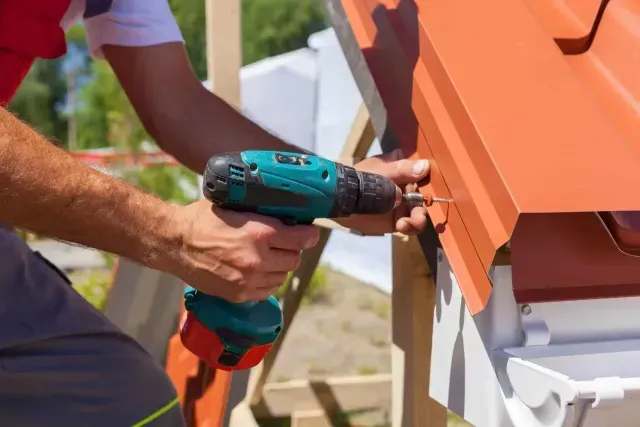 Roof repair services in Aurora, IL for fastening a piece of orange metal roofing near a white gutter on a construction site.