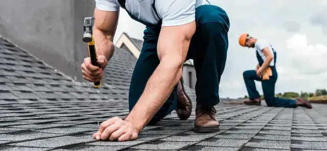 Two workers from a roof repair company in Aurora, IL, are kneeling on a residential roof, installing gray asphalt shingles with a hammer.