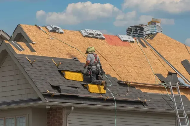 Roofing contractor in Aurora, IL installs shingles on a sloped residential roof under a bright blue sky.