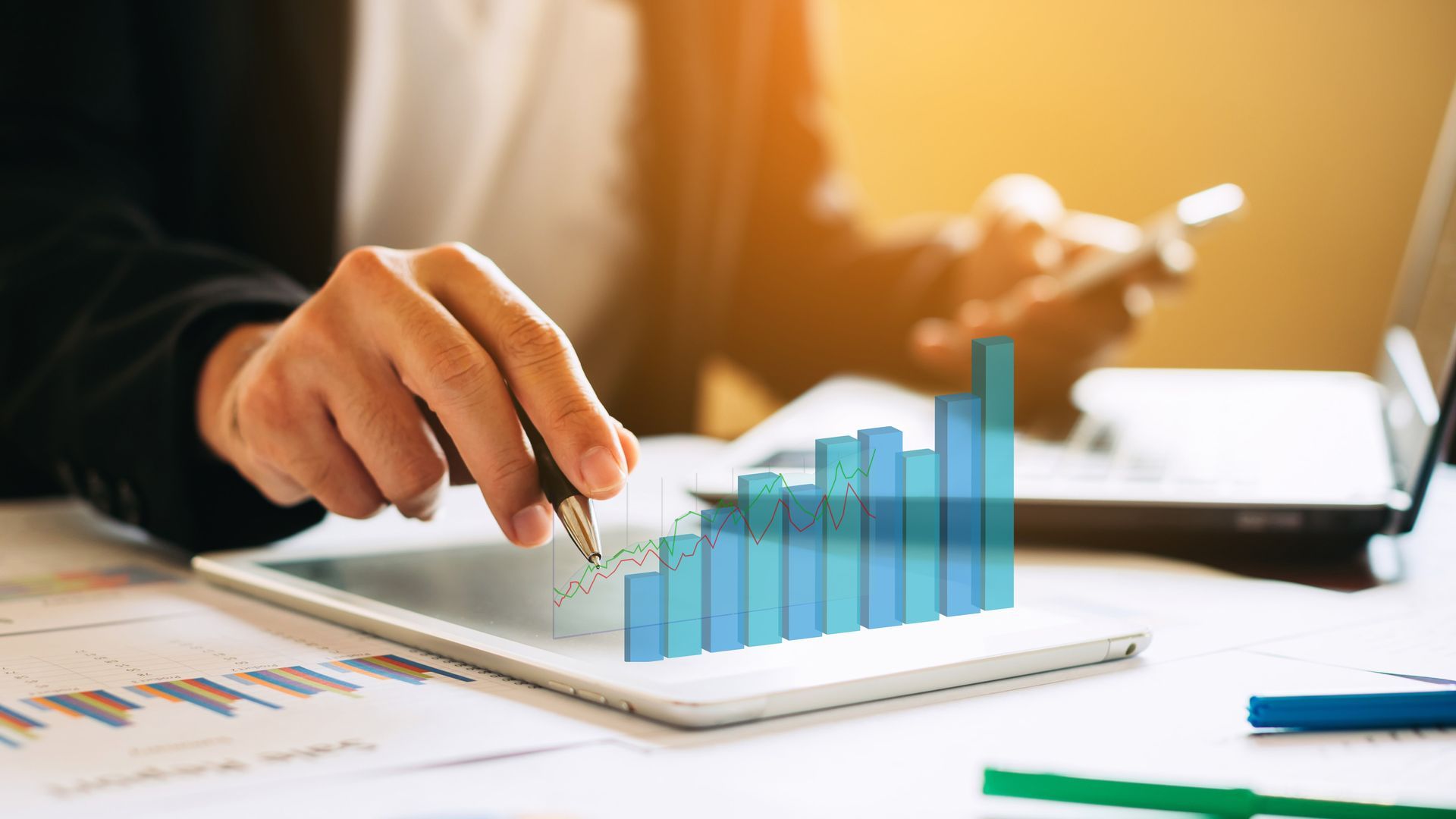 Person pointing to a growing bar graph on a tablet, with a laptop and phone in the background.
