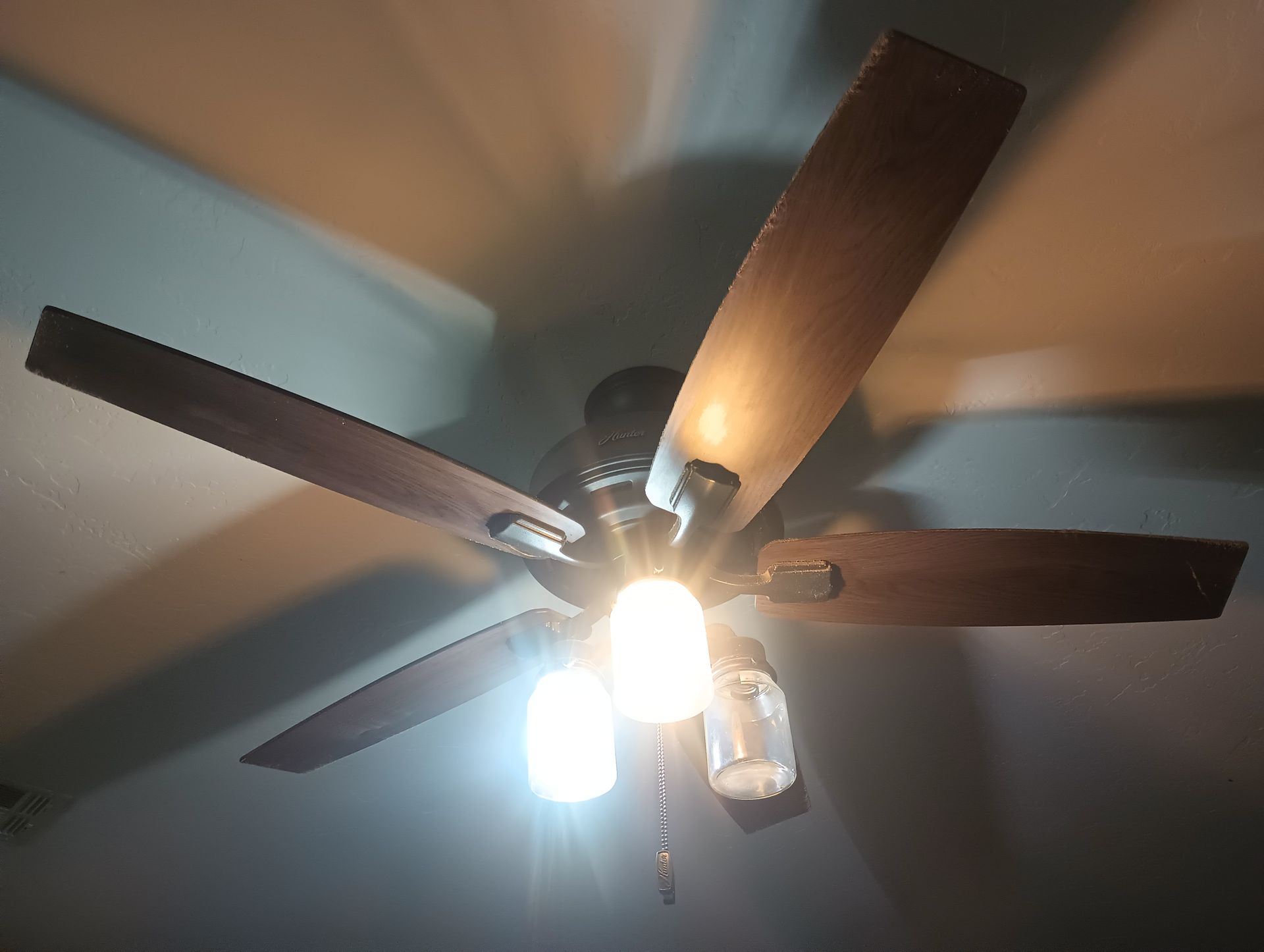 Ceiling fan with dark wooden blades and lit bulbs against a pale ceiling.