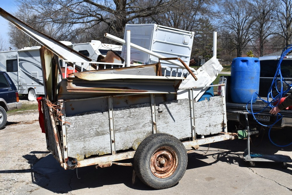 A trailer loaded with scrap metal and furniture sits outdoors beside blue barrels.