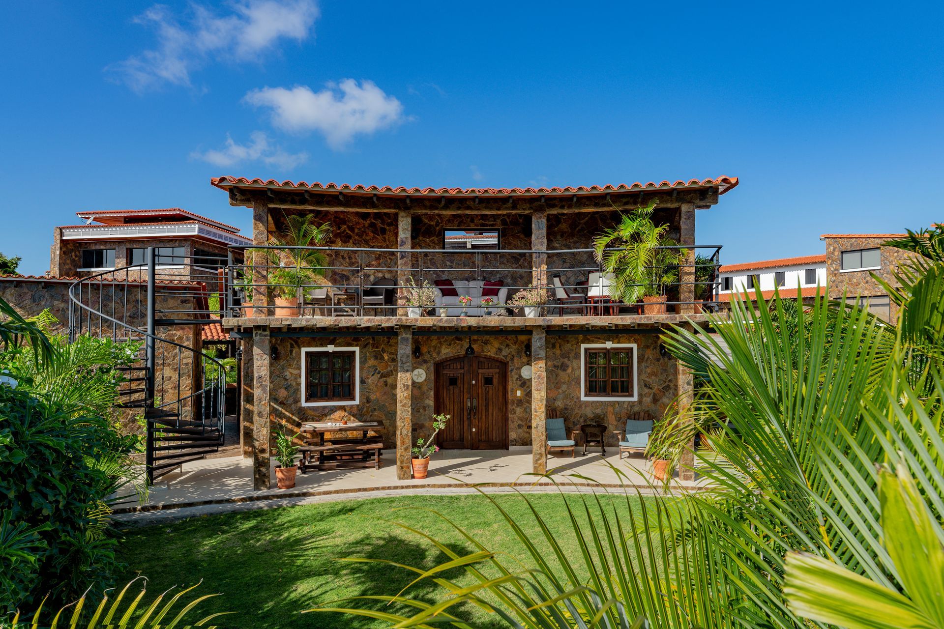 Stone two-story house with balcony, wooden doors, and lush tropical landscaping under a bright blue sky