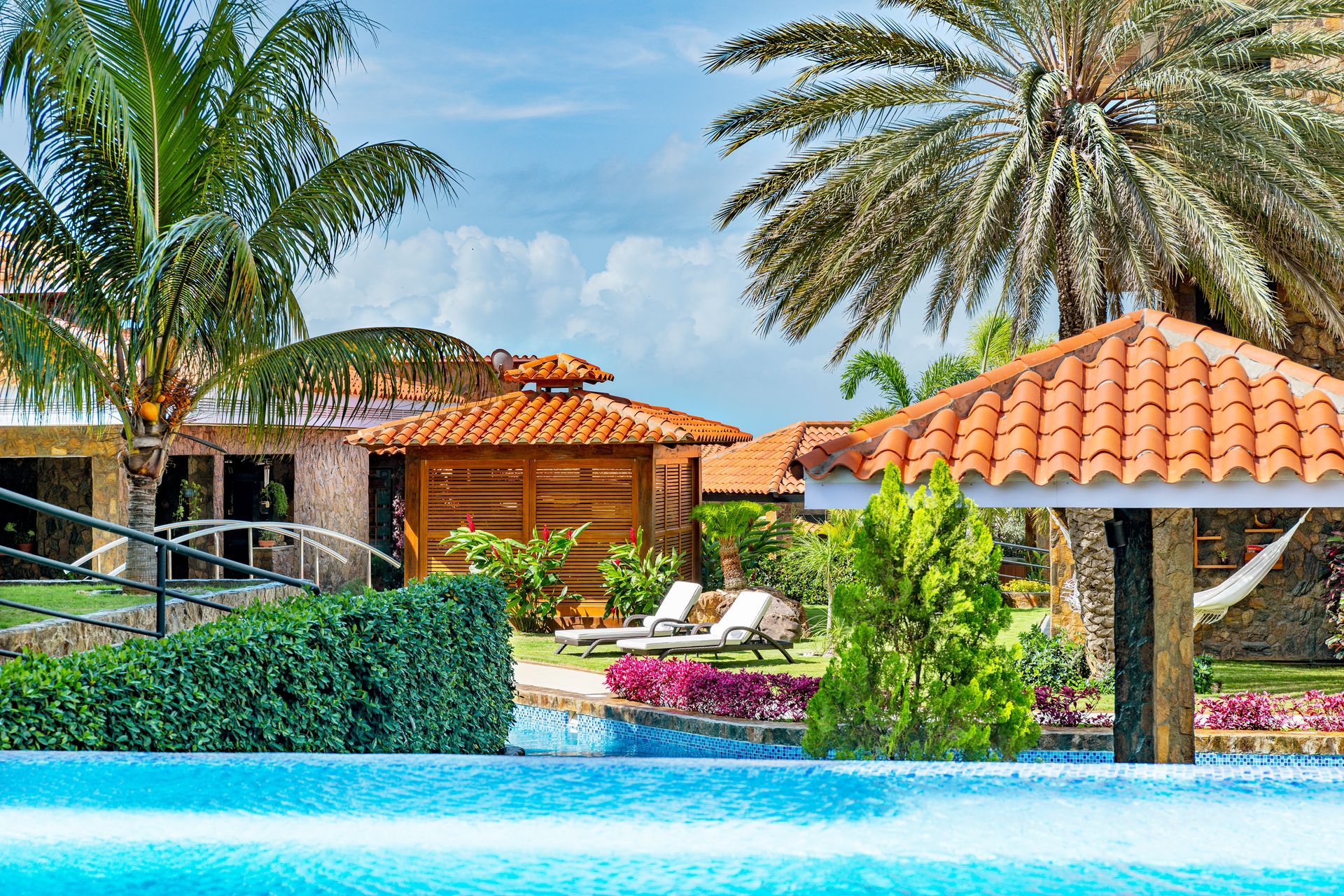 Tropical resort pool with palm trees, orange-tiled villas, and lounge chairs under a bright blue sky