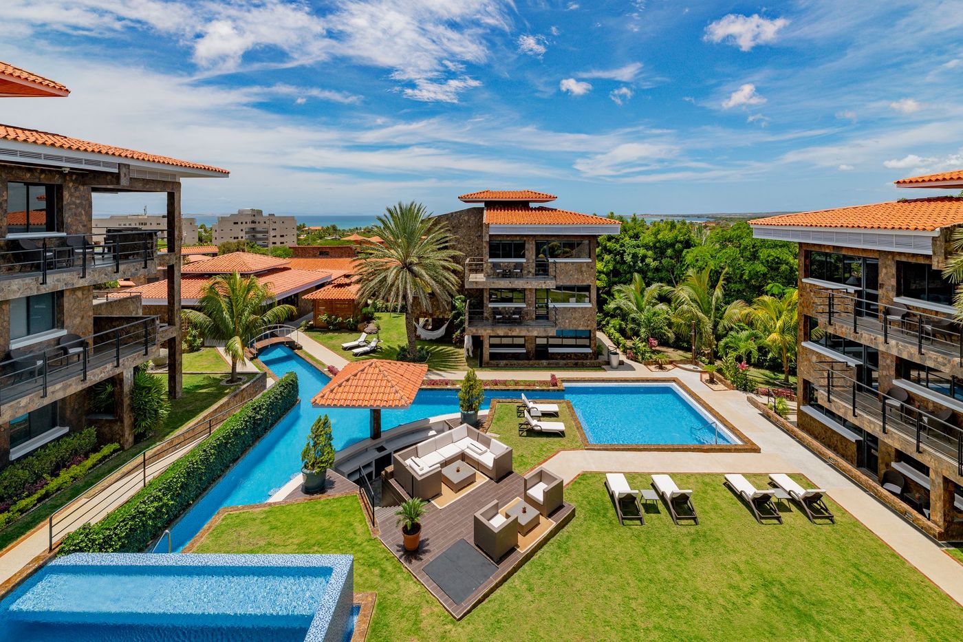 Resort courtyard with pool, lounge chairs, palm trees, and terracotta-roofed buildings under a blue sky