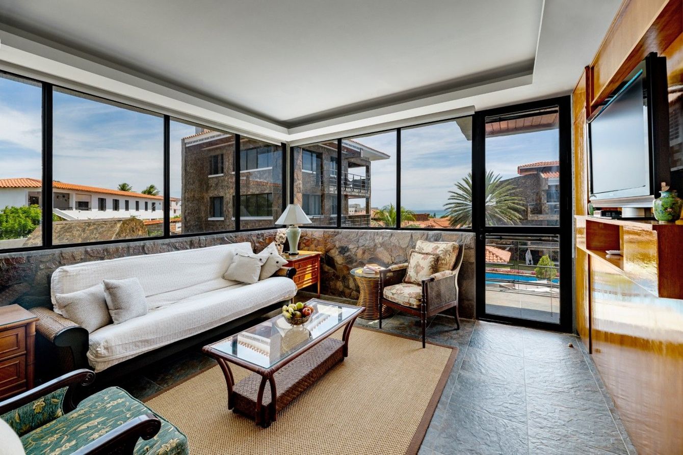 Bright living room with white sofa, wicker chairs, and large windows overlooking a balcony and city view