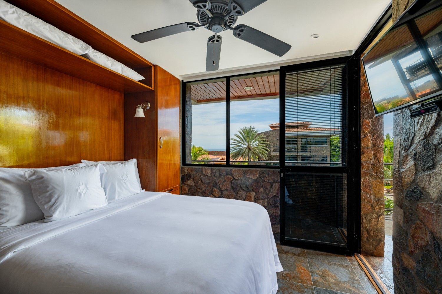 Modern bedroom with white bed, wood paneling, ceiling fan, and balcony view through large windows