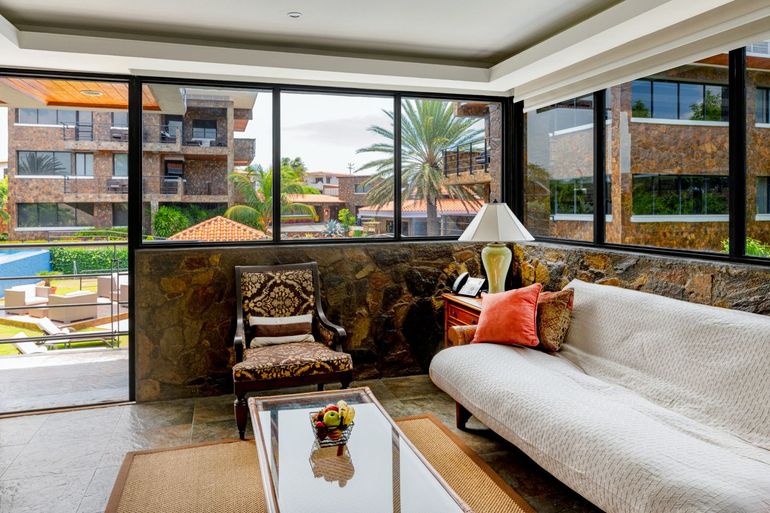 Modern living room with white sofa, patterned chair, glass coffee table, and open view to a balcony and apartments