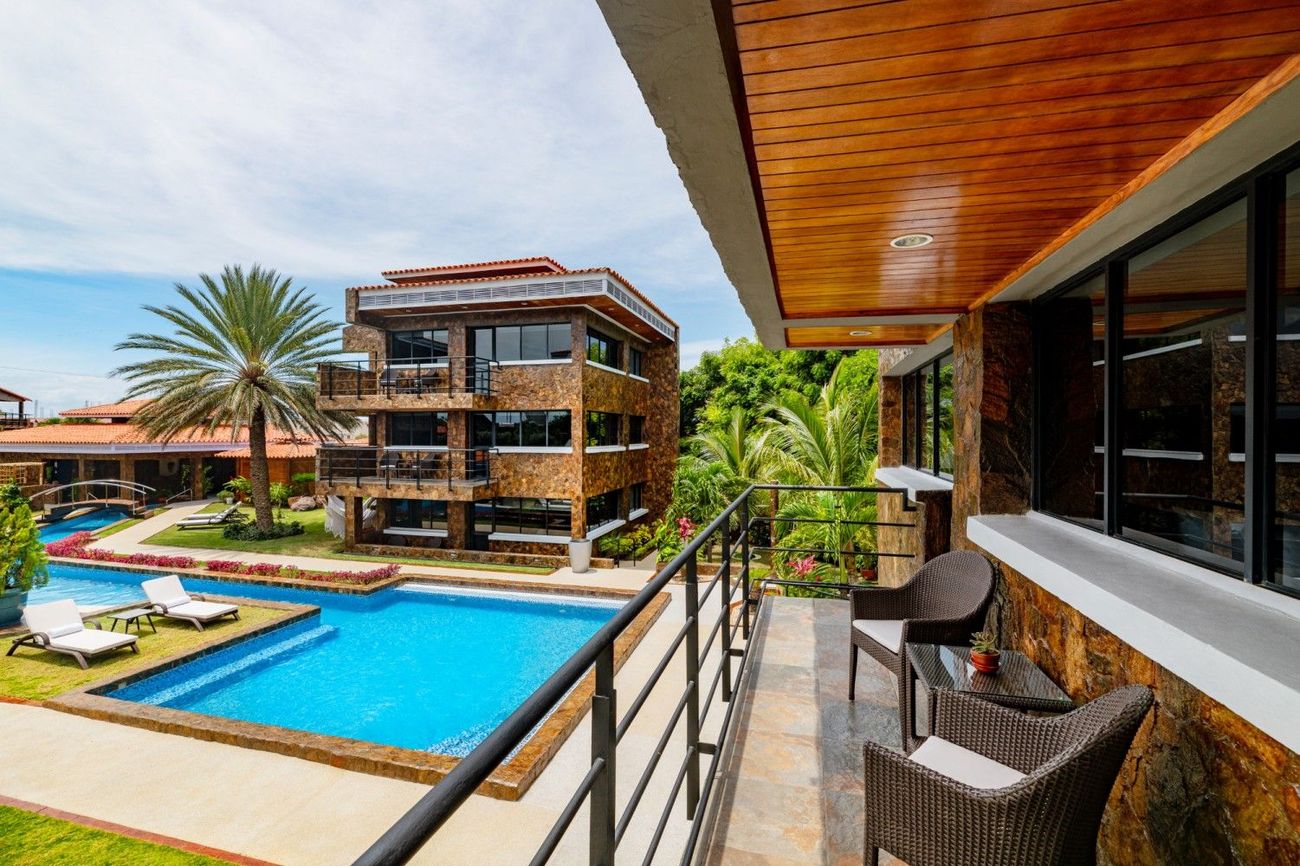 Resort balcony overlooking a turquoise pool, palm trees, and modern villas under a wooden canopy