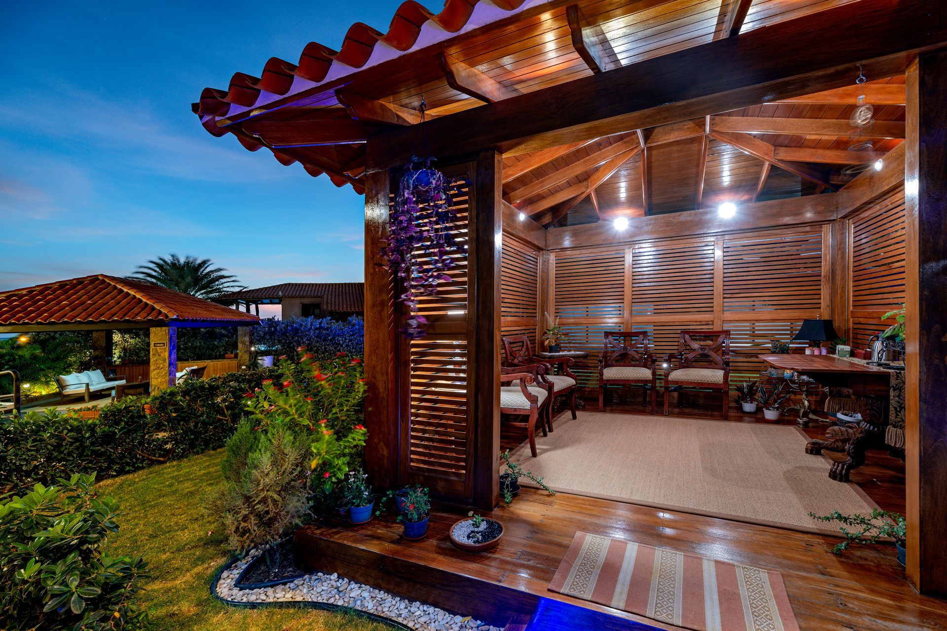 Rustic open-air patio at dusk with warm lights, wooden beams, woven furniture, and garden landscaping