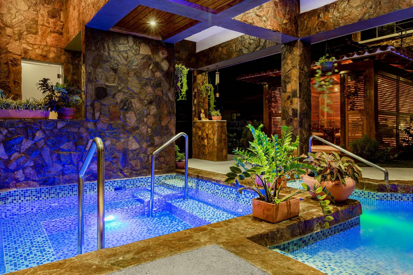 Luxury outdoor pool and hot tub lit blue and green beside a stone patio and tropical plants at night