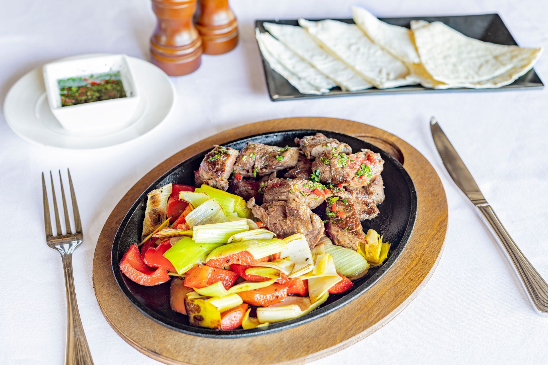 Sizzling steak and vegetables in a cast-iron skillet with tortillas and dipping sauce on a white table