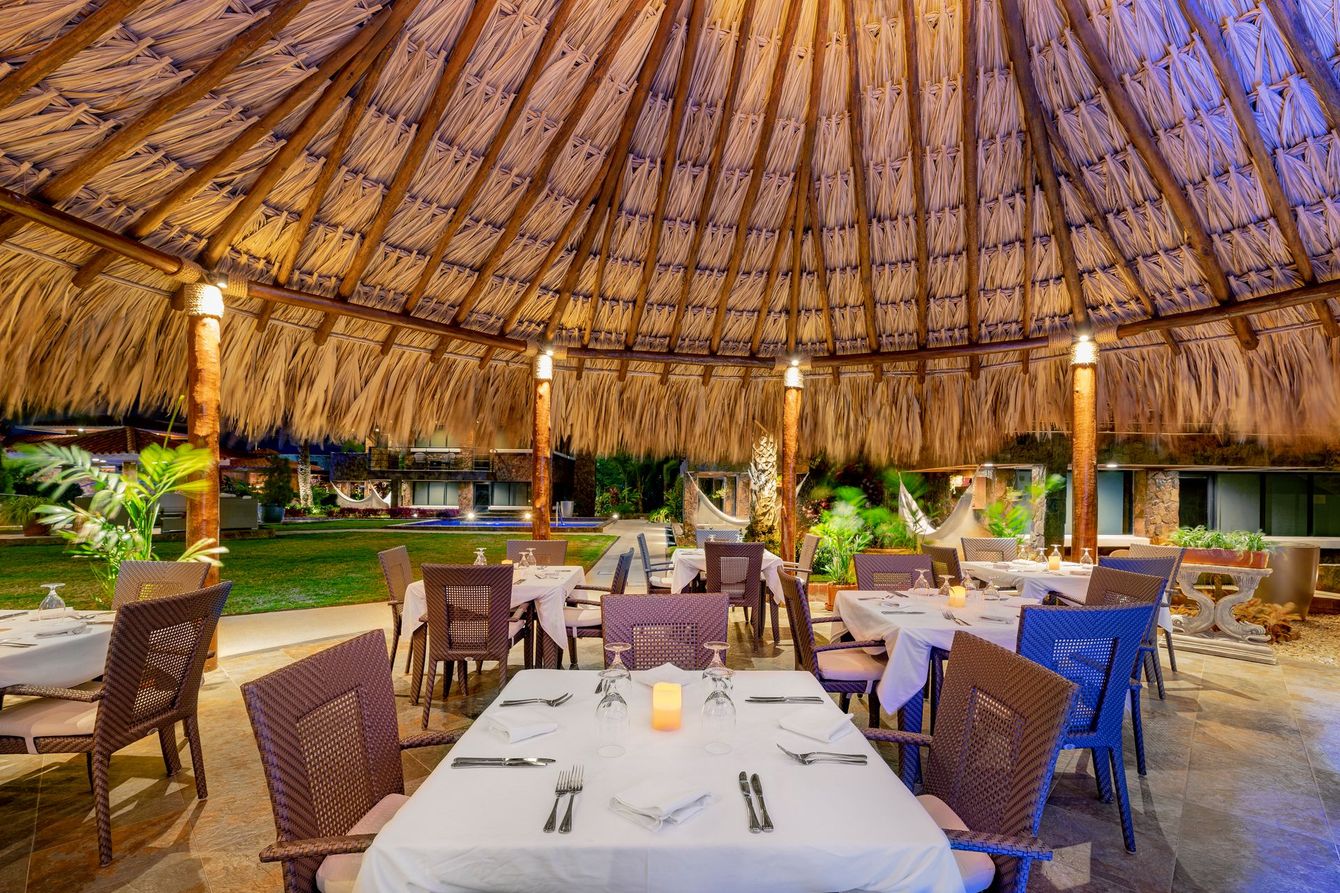 Open-air thatched restaurant with white tablecloths and woven chairs at dusk