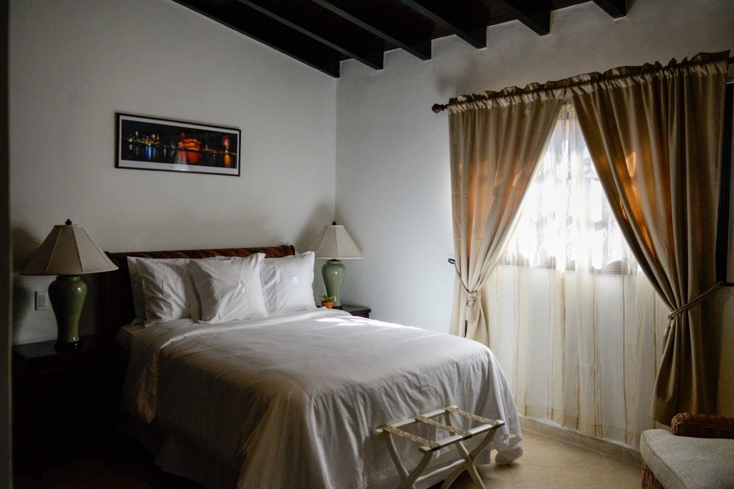 Cozy bedroom with a white bed, dark wood beams, and sunlight through sheer curtains.