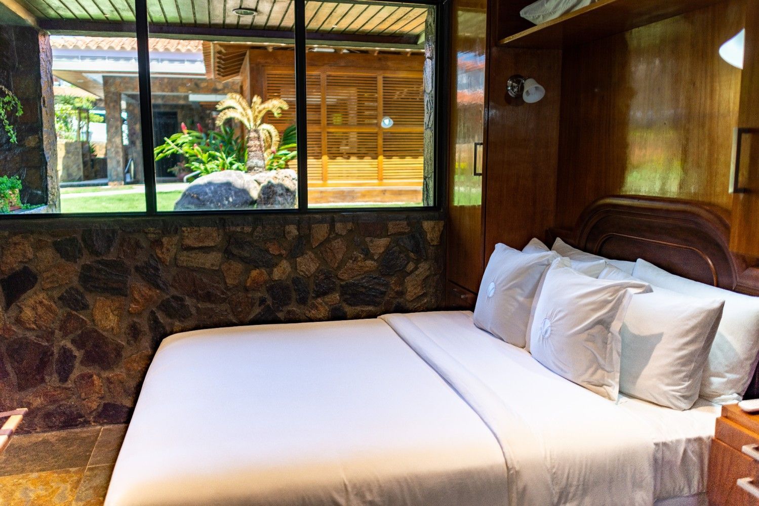 Cozy bedroom with white bed linens, wood furniture, stone wall, and window view of a garden patio