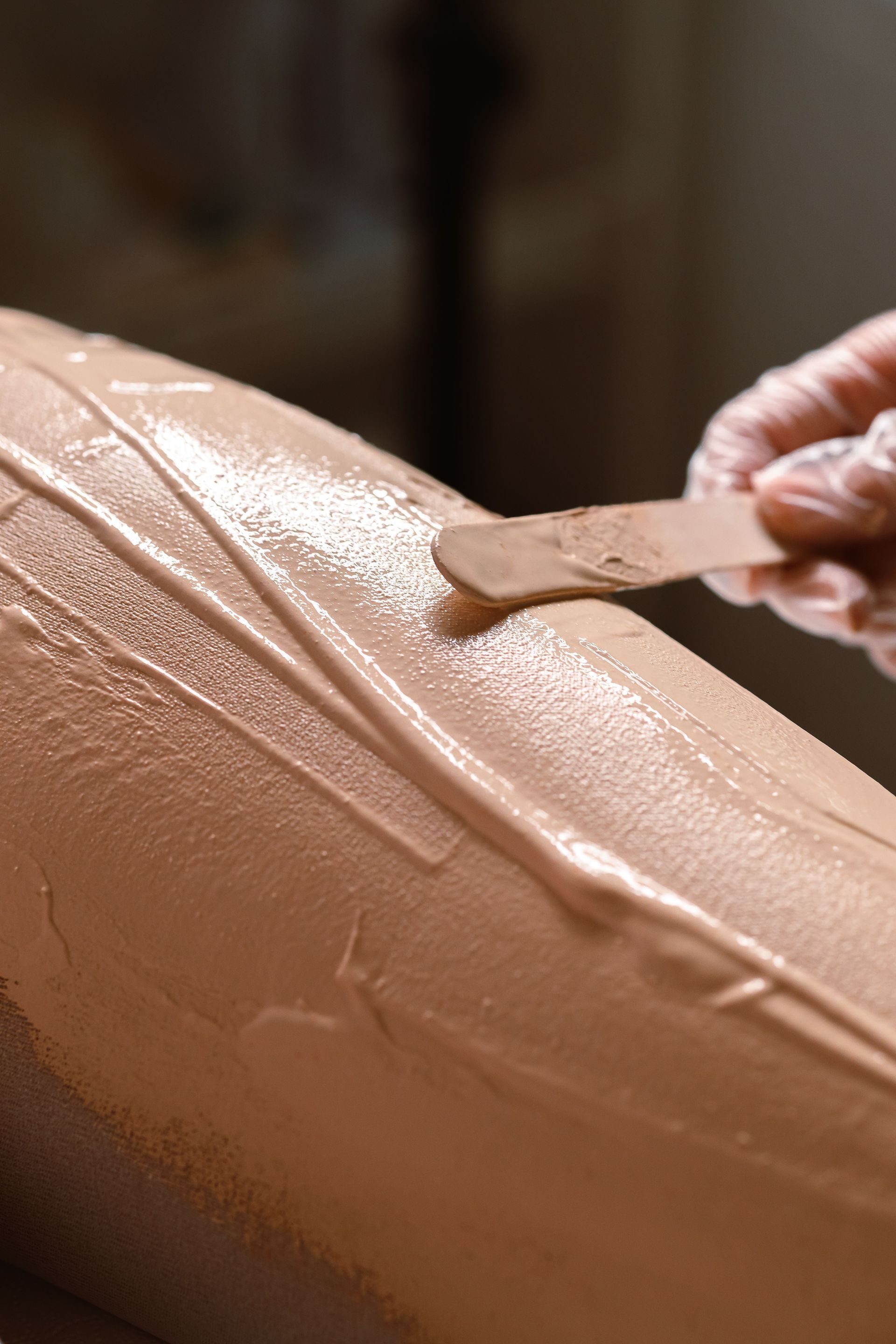 A hand spreads beige cream onto skin with a wooden spatula.