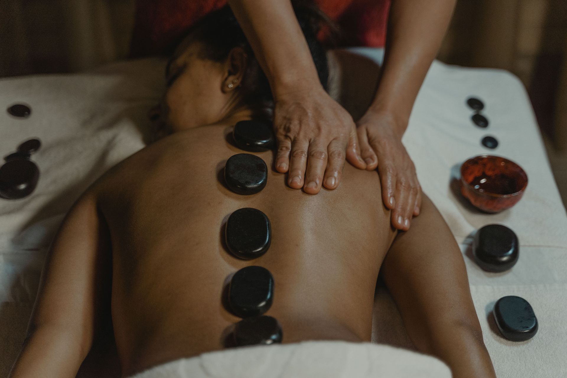 Person receiving a hot stone massage on a table, with stones aligned along the back and hands massaging shoulders.