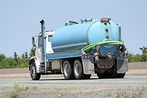 Blue vacuum tanker truck on a road, with hoses attached at the rear under a clear sky