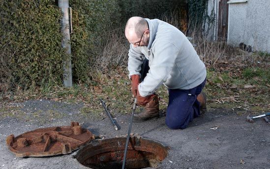 Worker in gray sweatshirt kneels by an open manhole, using tools beside a rusted cover.
