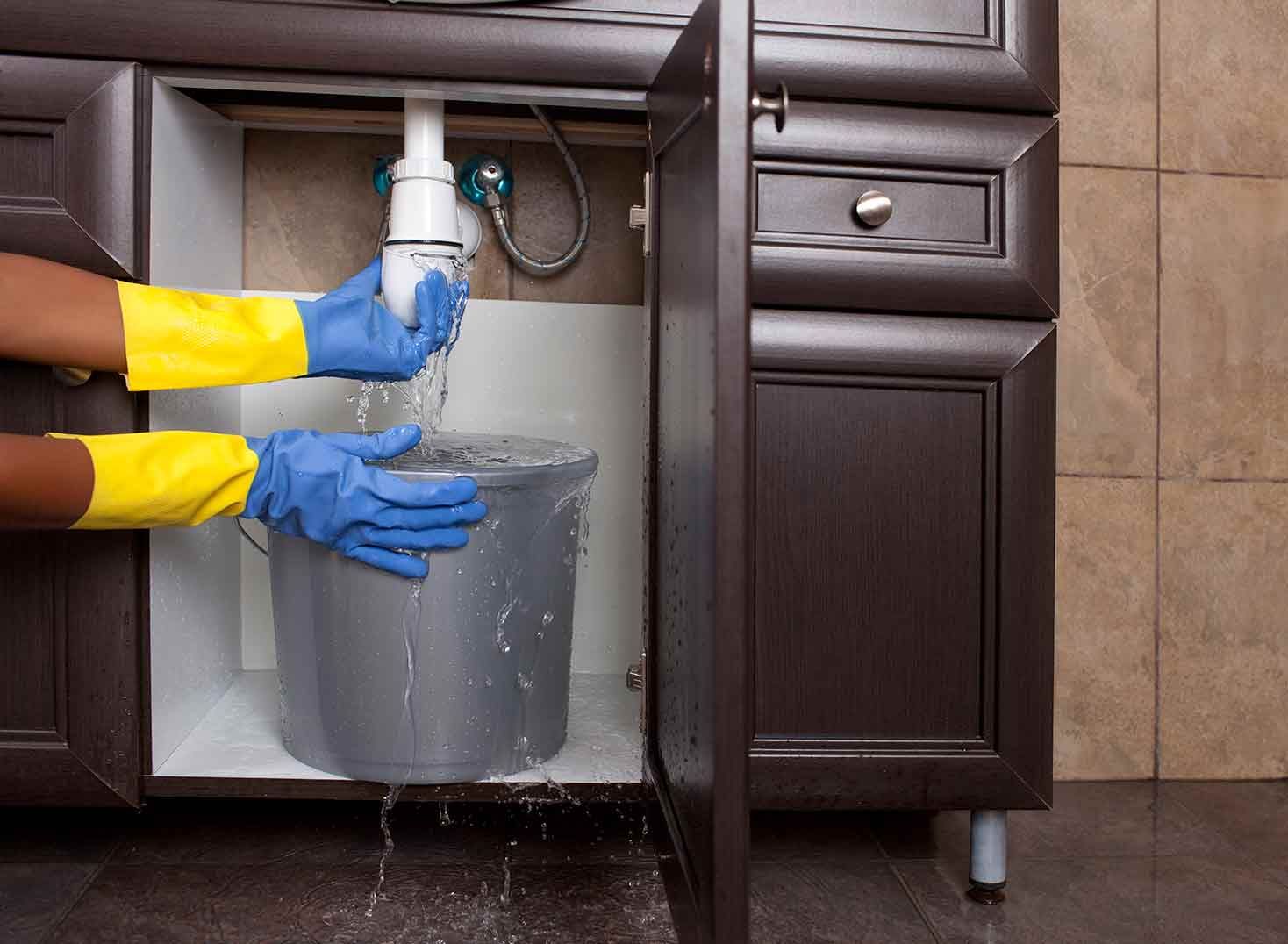 Plumber in yellow sleeves fixing a leaky pipe under a dark bathroom sink, water dripping into a bucket.