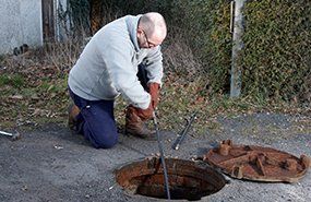 Worker using a long tool to open a round manhole cover on a road and inspect the opening