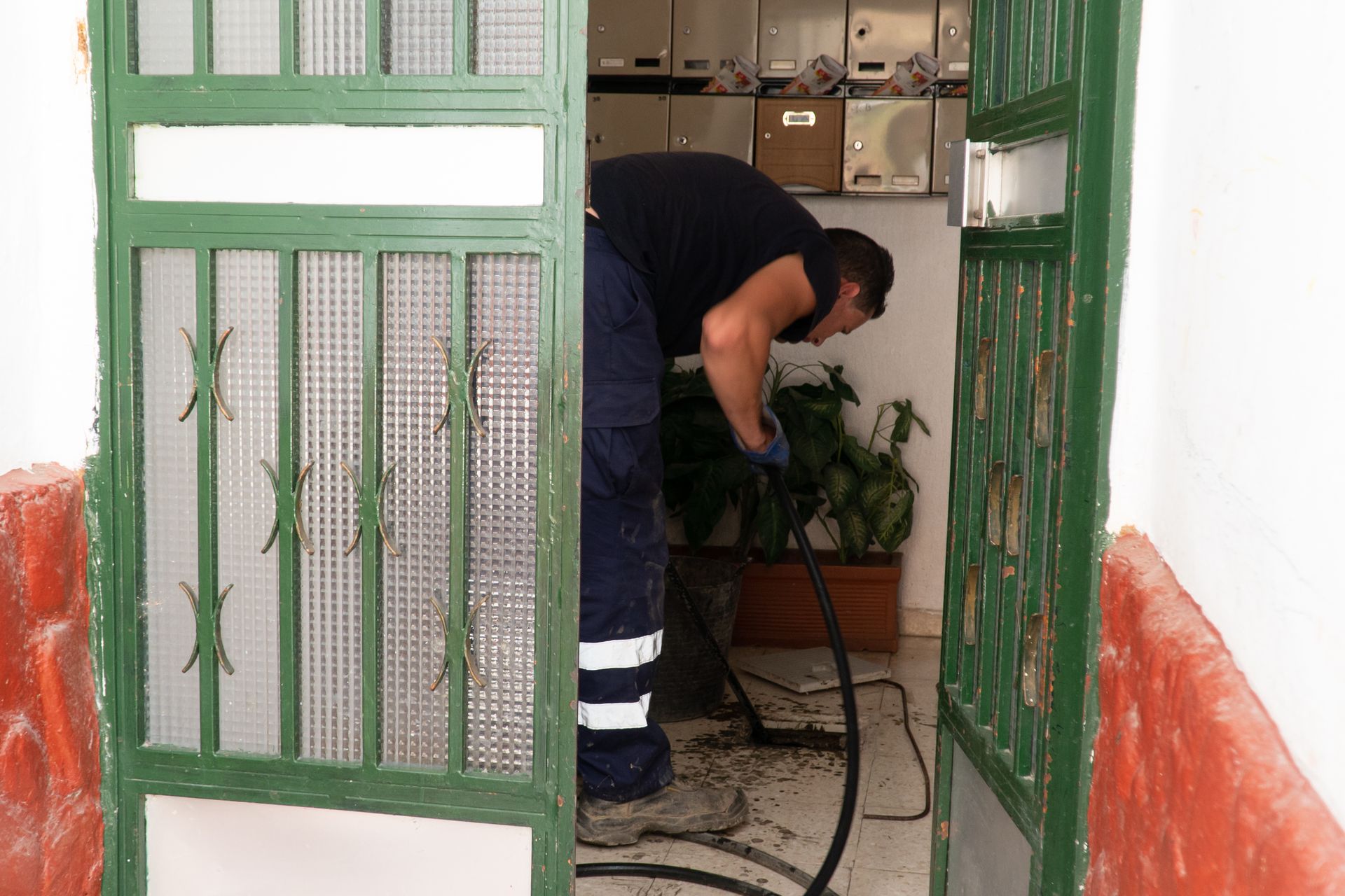Person bending to water a potted plant by a green metal gate indoors