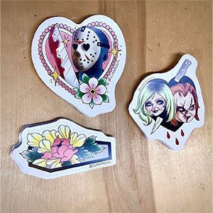 Three stickers are sitting on a wooden table including jason voorhees and chucky