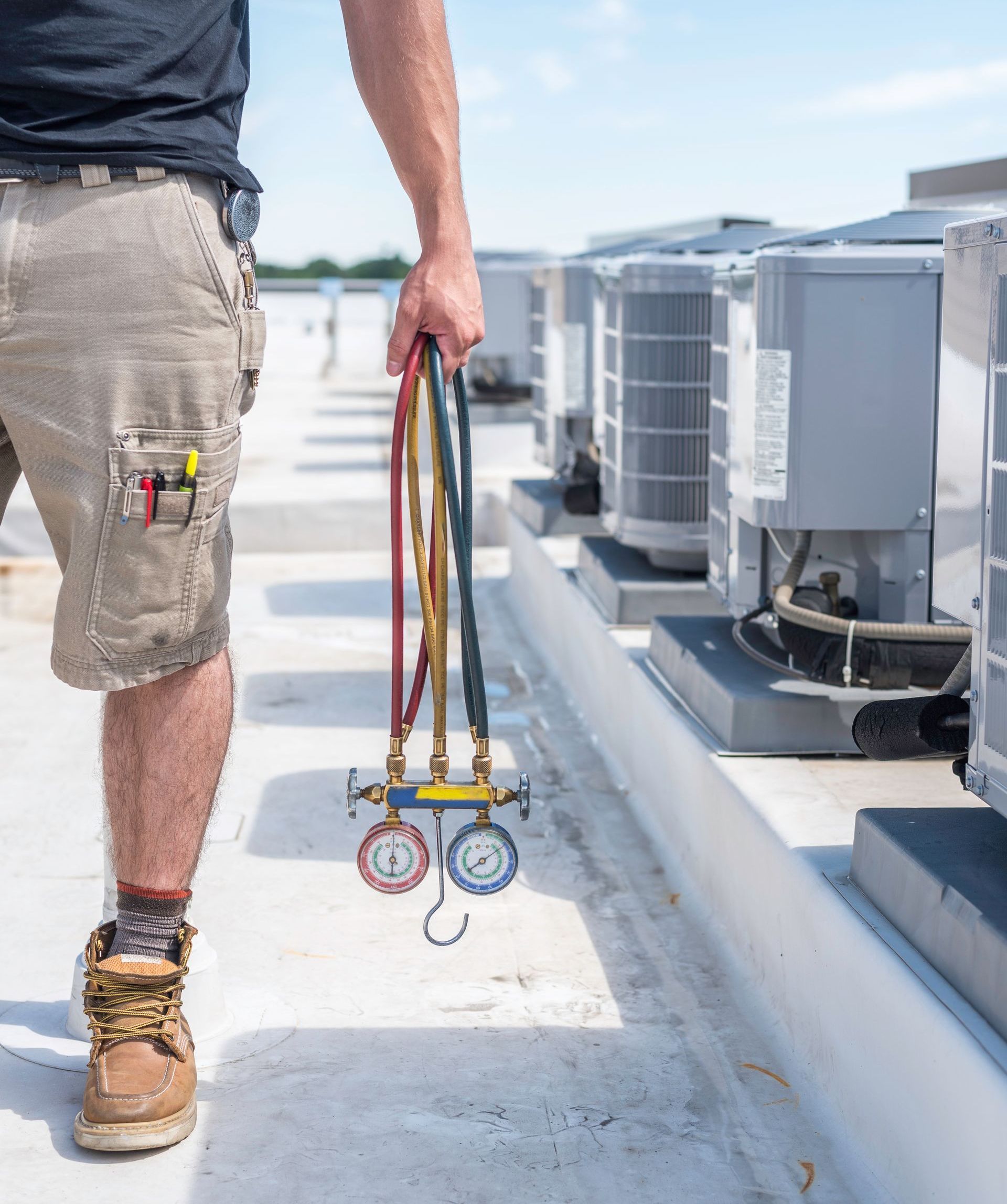 HVAC technician holding gauges on a rooftop next to several air conditioning units.