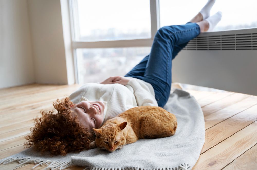 Woman lying on floor with cat, smiling. Legs up on windowsill.