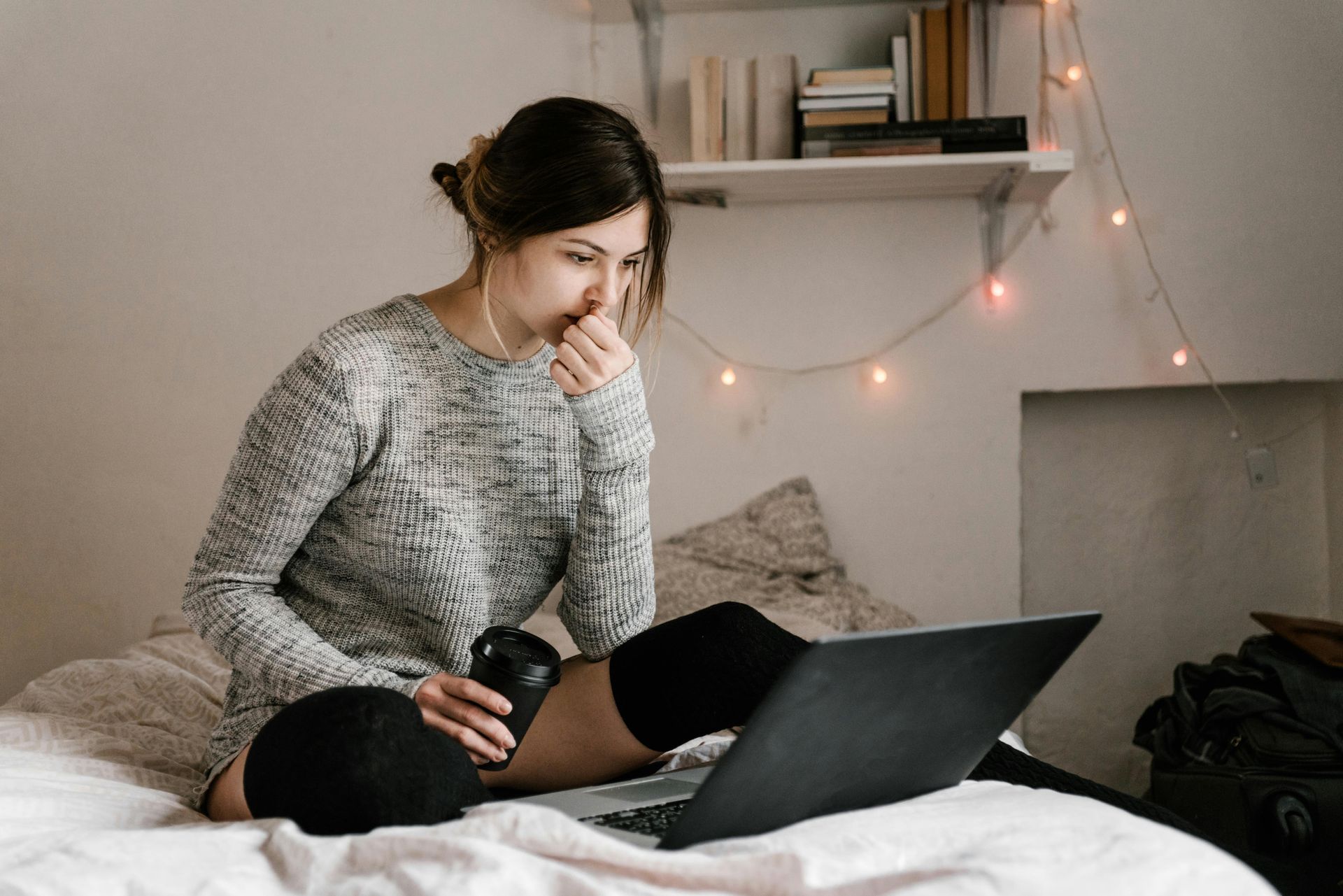 Woman on bed using laptop, holding coffee, thoughtful expression, in a cozy room.