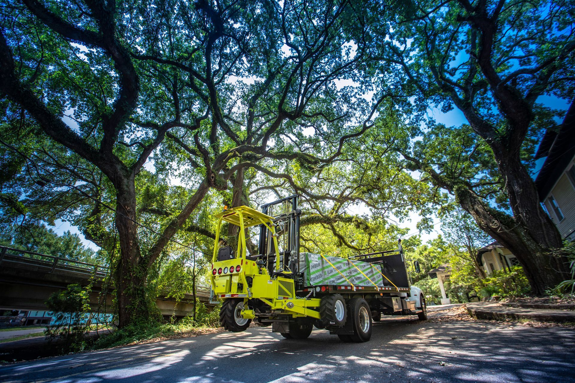 Yellow forklift lifting cargo on a truck, parked under large trees.