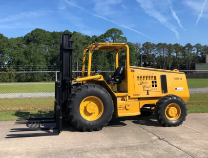 Yellow construction forklift parked on a concrete lot with trees and a fence in the background
