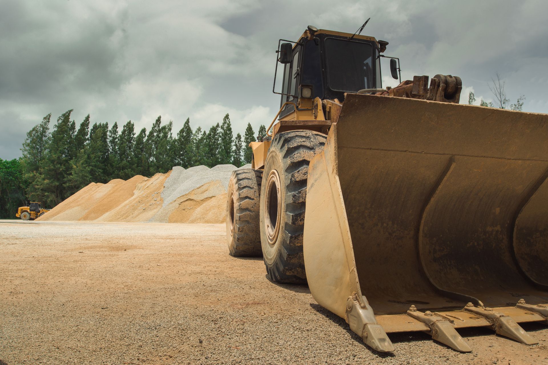 Bulldozer with raised blade on a gravel site beside sand piles and trees