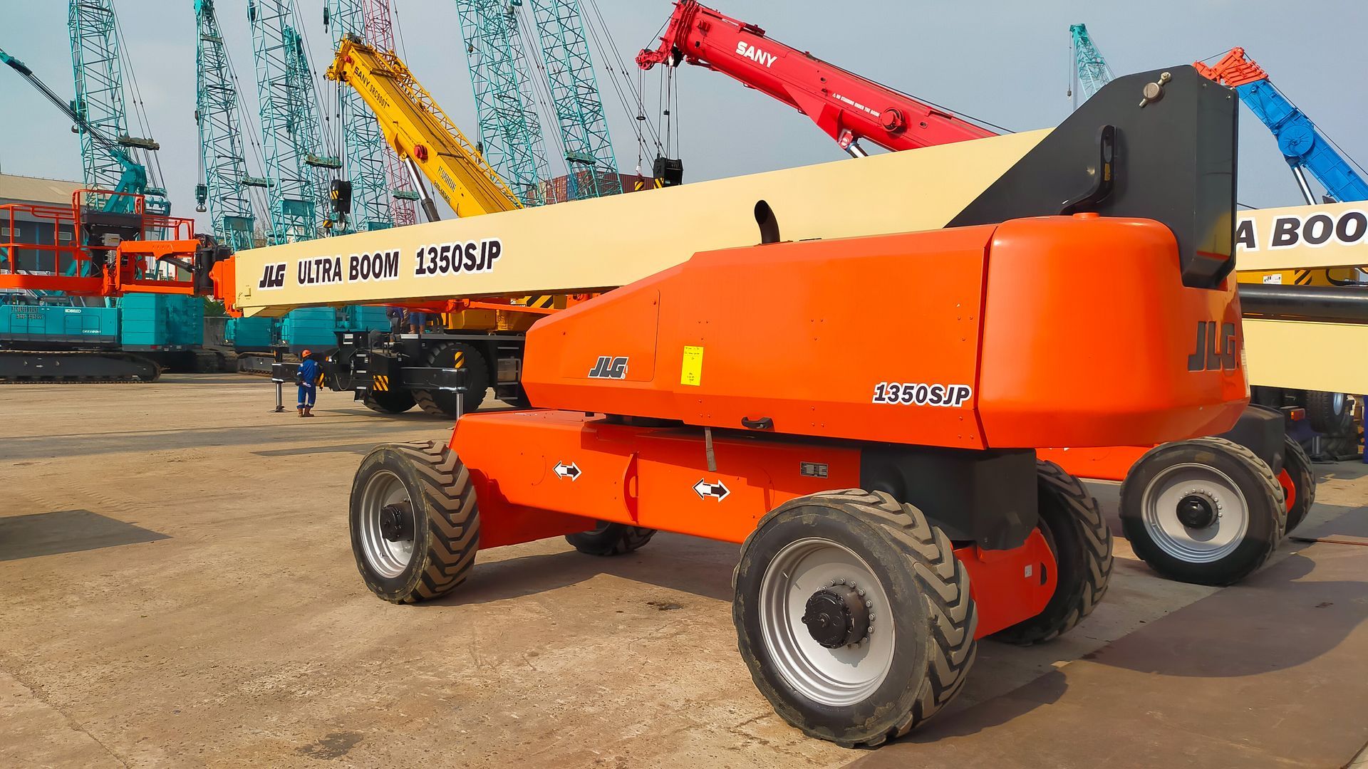 Orange boom lift parked outdoors at a construction equipment yard, with other cranes in the background