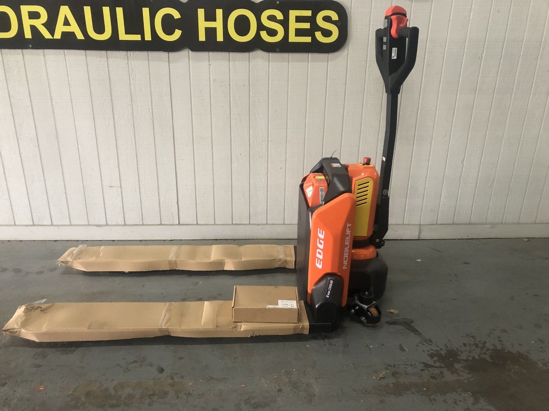 Orange pallet jack with two wrapped pallets in a warehouse under a hydraulic hoses sign