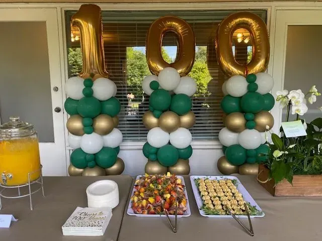 100th birthday party setup with gold balloon numbers, green-white balloon columns, and dessert table
