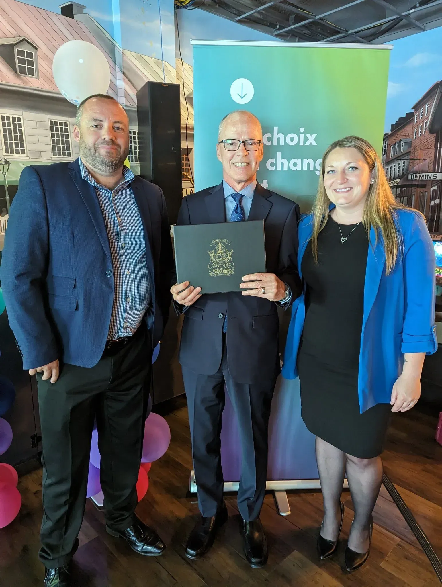Three people standing indoors at an award presentation, holding a certificate in front of a blue-green banner.