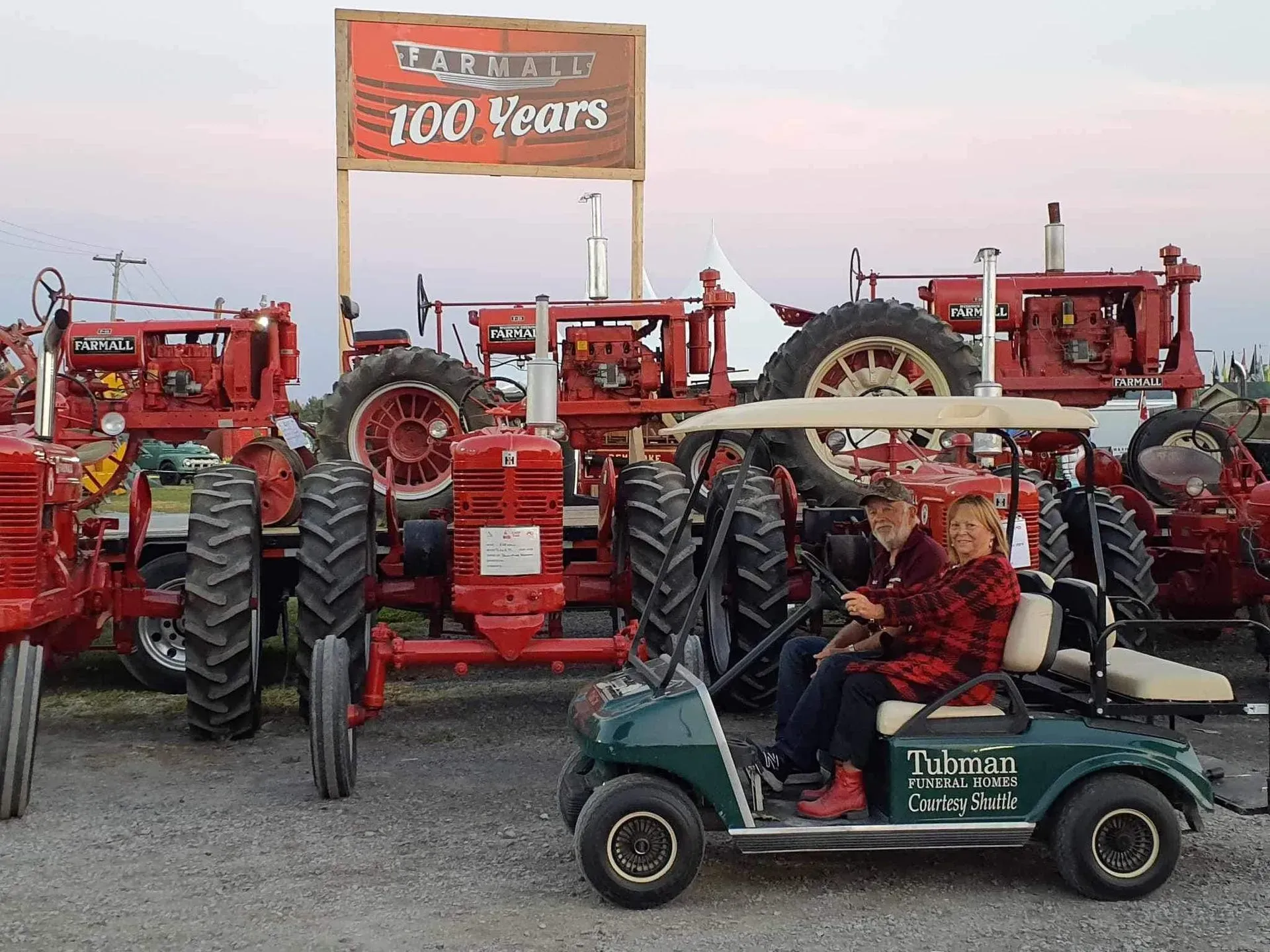 Vintage red tractors lined up at a fair, with a sign reading “100 Years” and a golf cart in front