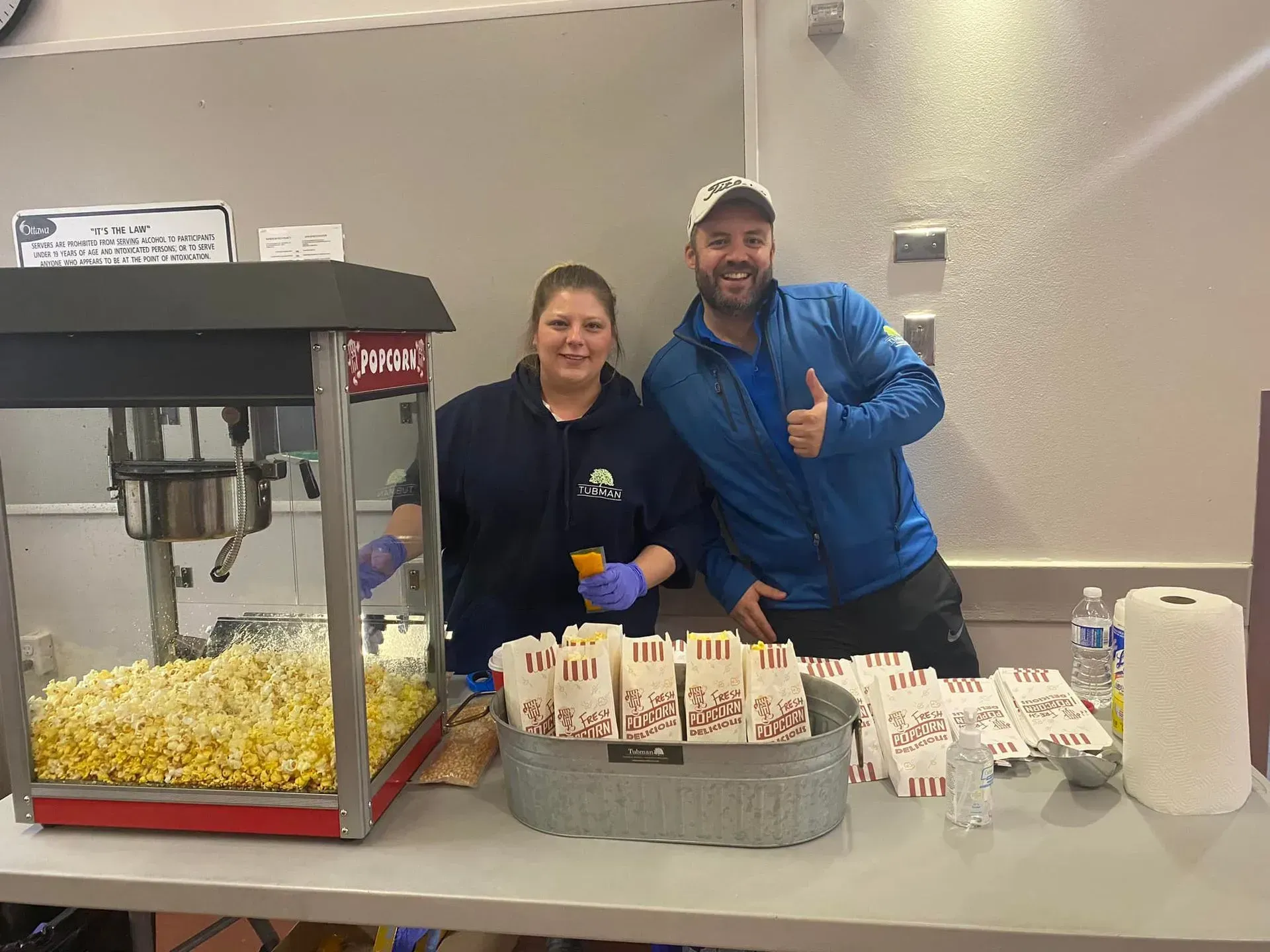 Two people at a concession table with popcorn, drinks, and snacks, smiling and giving thumbs up.