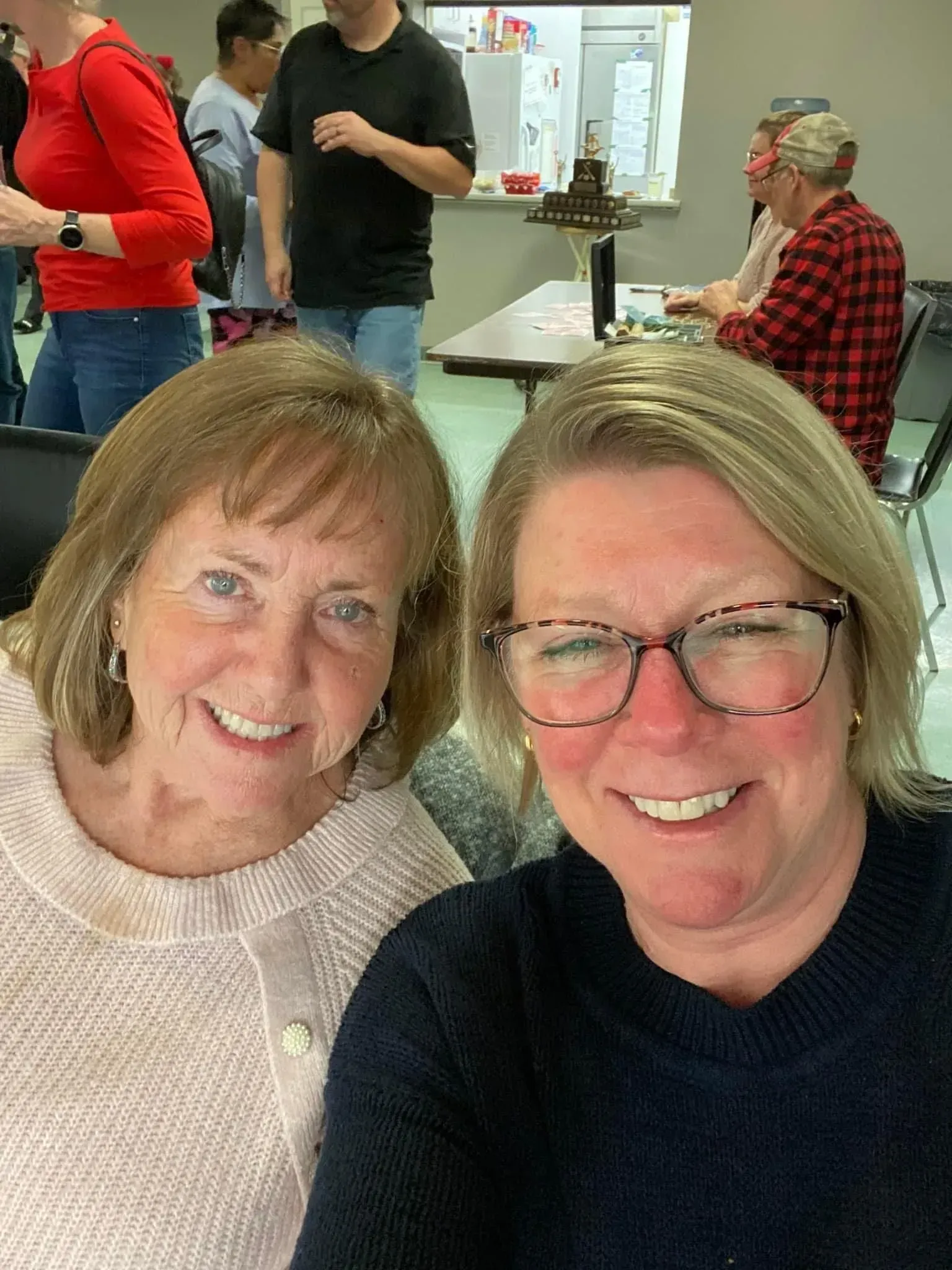 Two smiling people pose indoors at a casual gathering, with others and a table in the background.