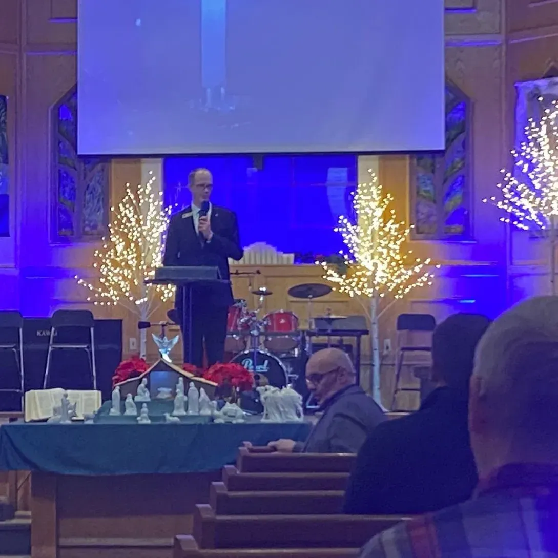 Speaker at podium on a blue-lit stage with lit trees and a drum set in the background