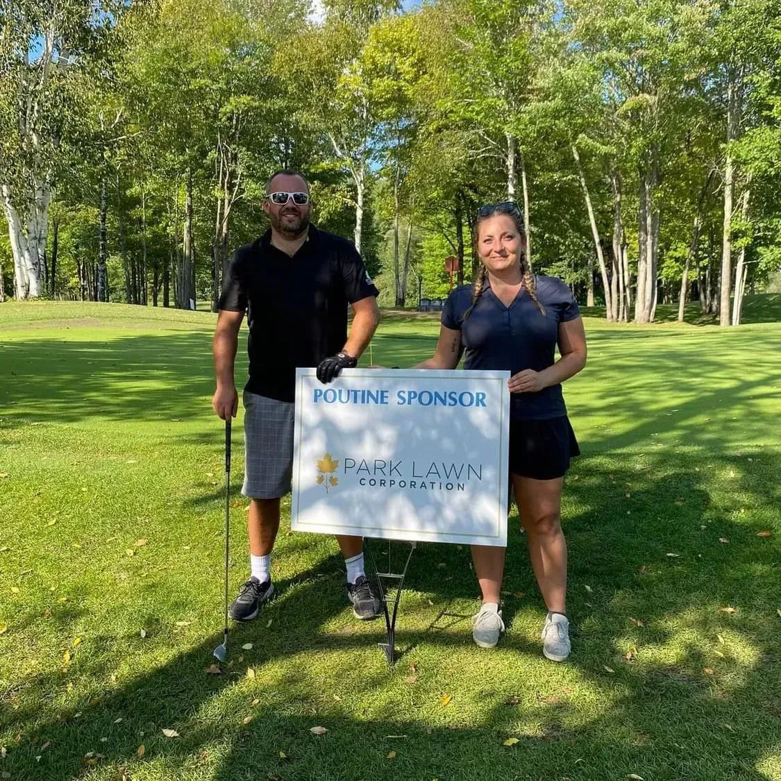 Two golfers standing on a sunny fairway holding a white sponsor sign in front of trees