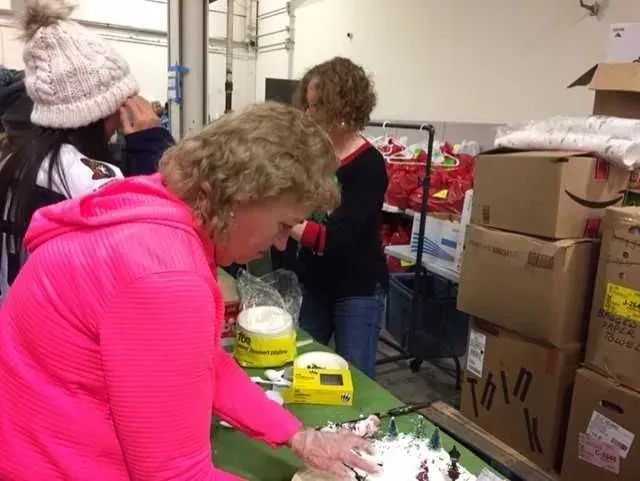 People packing supplies at a table in a storage room with stacked boxes and bins.