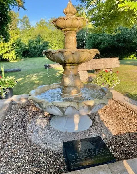 Stone tiered fountain in a garden, with water flowing into a gravel basin and greenery around it