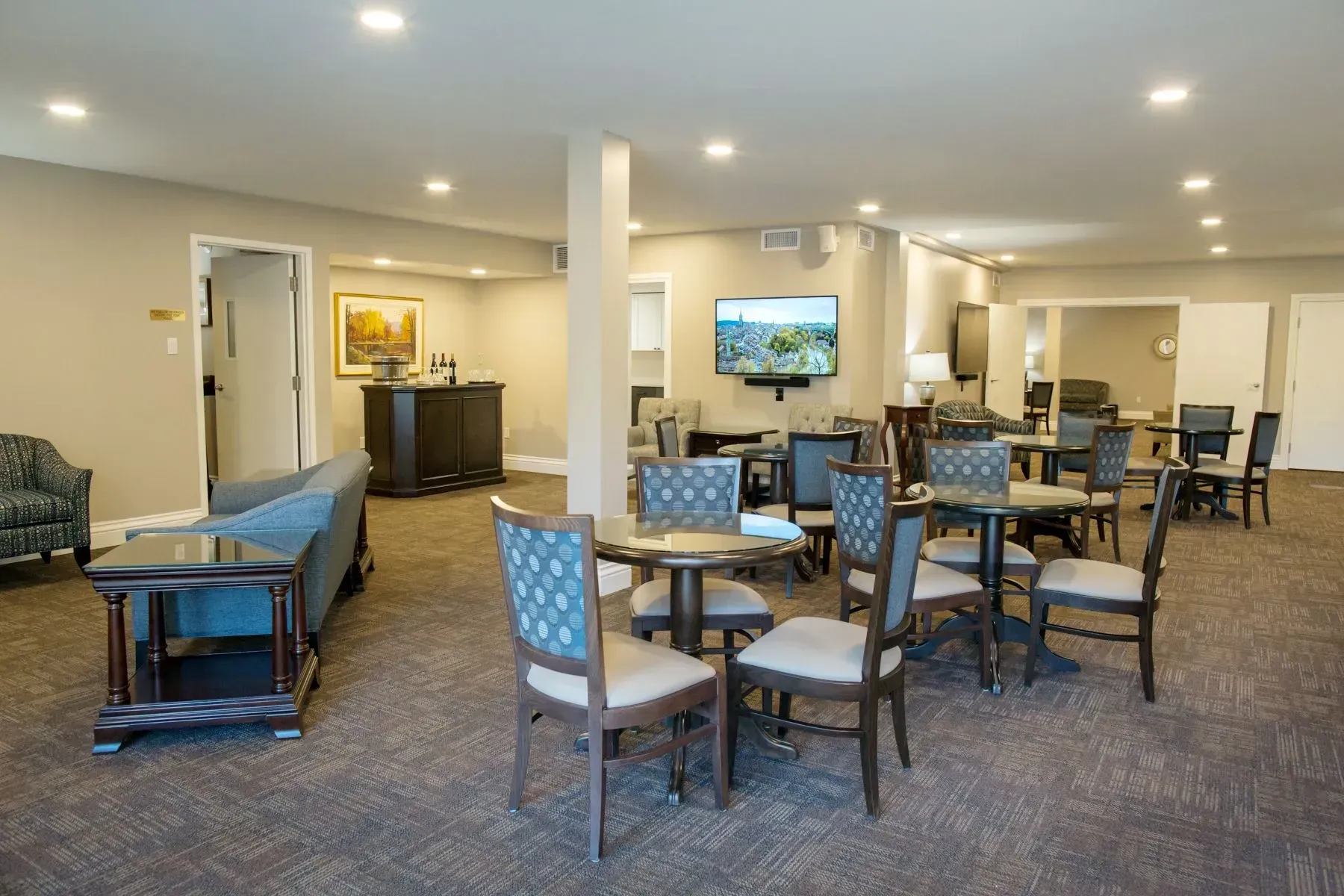 Spacious lounge with chairs, tables, couches, and a wall-mounted TV under recessed lights.