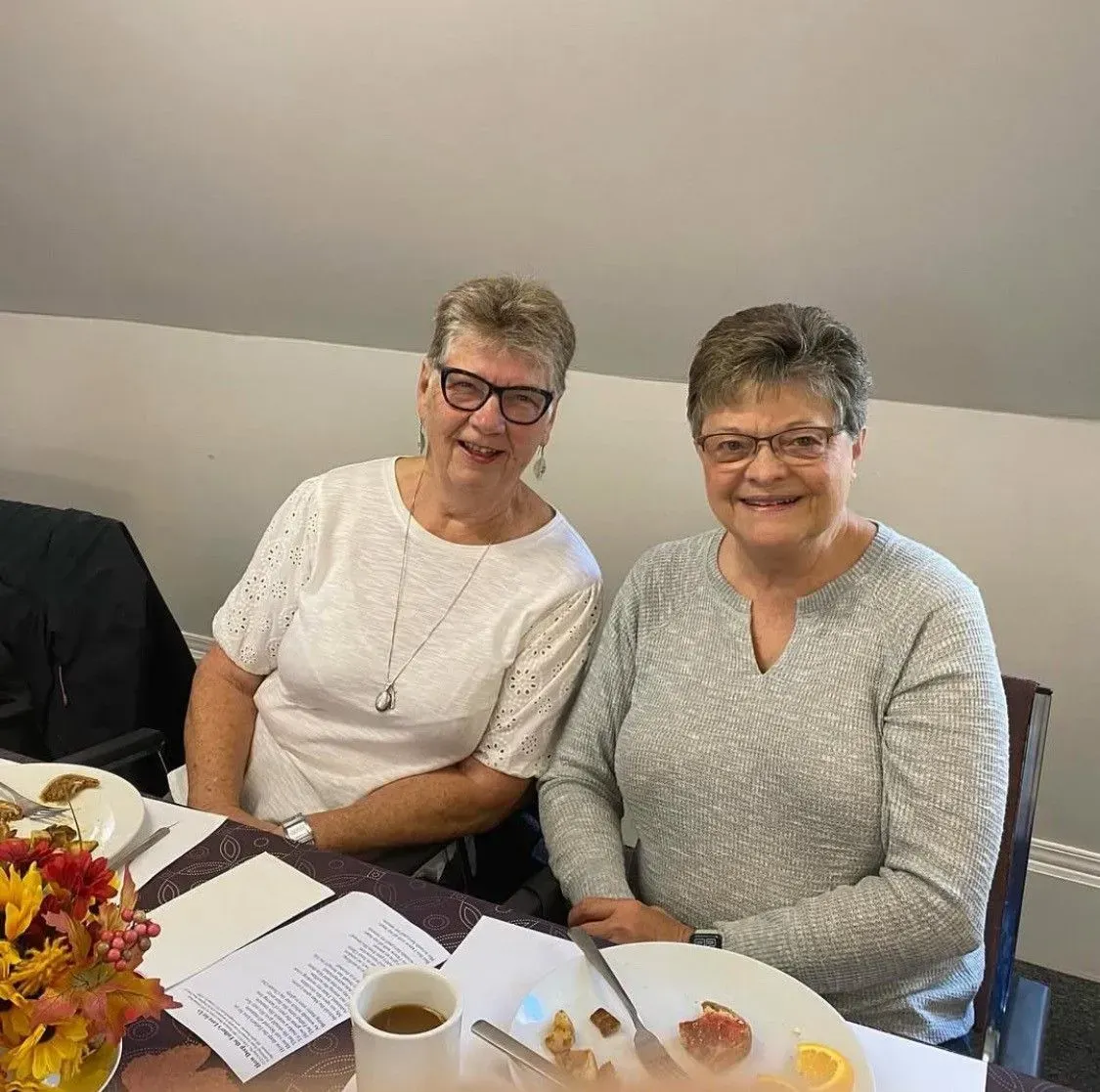 Two smiling people seated at a table with coffee, pastries, and flowers in a bright room