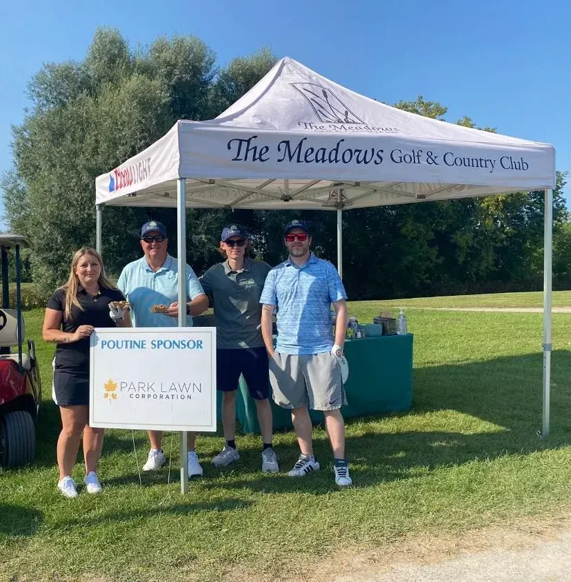 Four people posing under a Meadows Golf & Country Club tent beside a sponsor sign on a sunny lawn