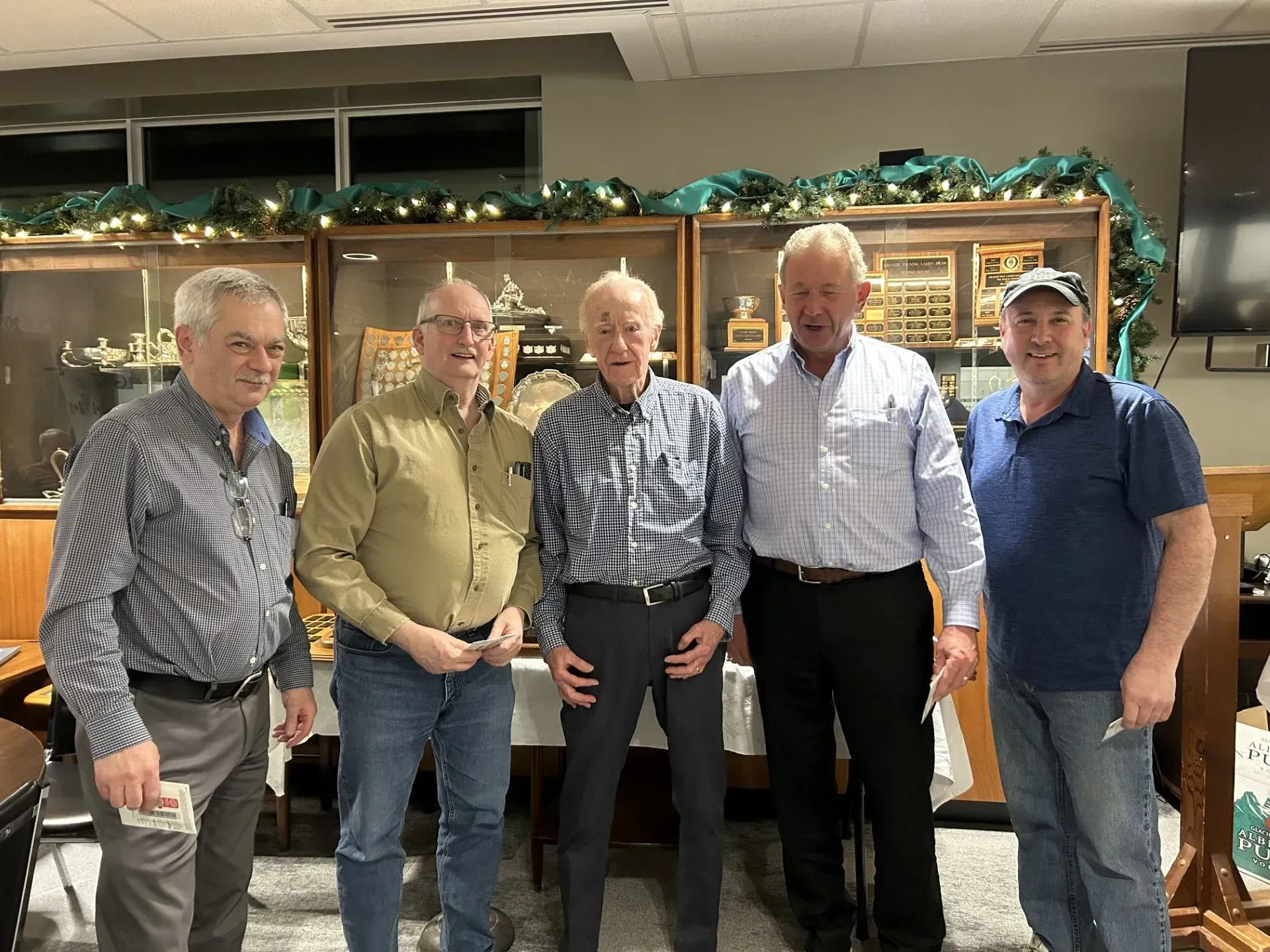 Five men standing together indoors, smiling in front of garland-decorated shelving.