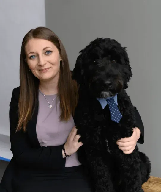 Woman kneels beside a black dog in a blue tie against a gray backdrop.